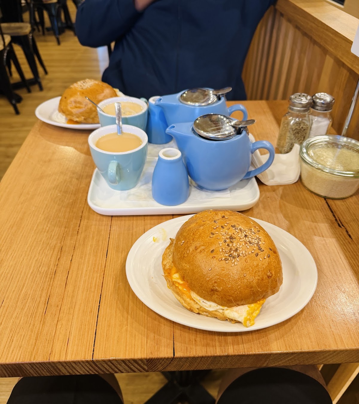 Blue teapots and cups on a tray beside a sesame seed roll sandwich on a wooden table at Salt n Pepa Cafe.