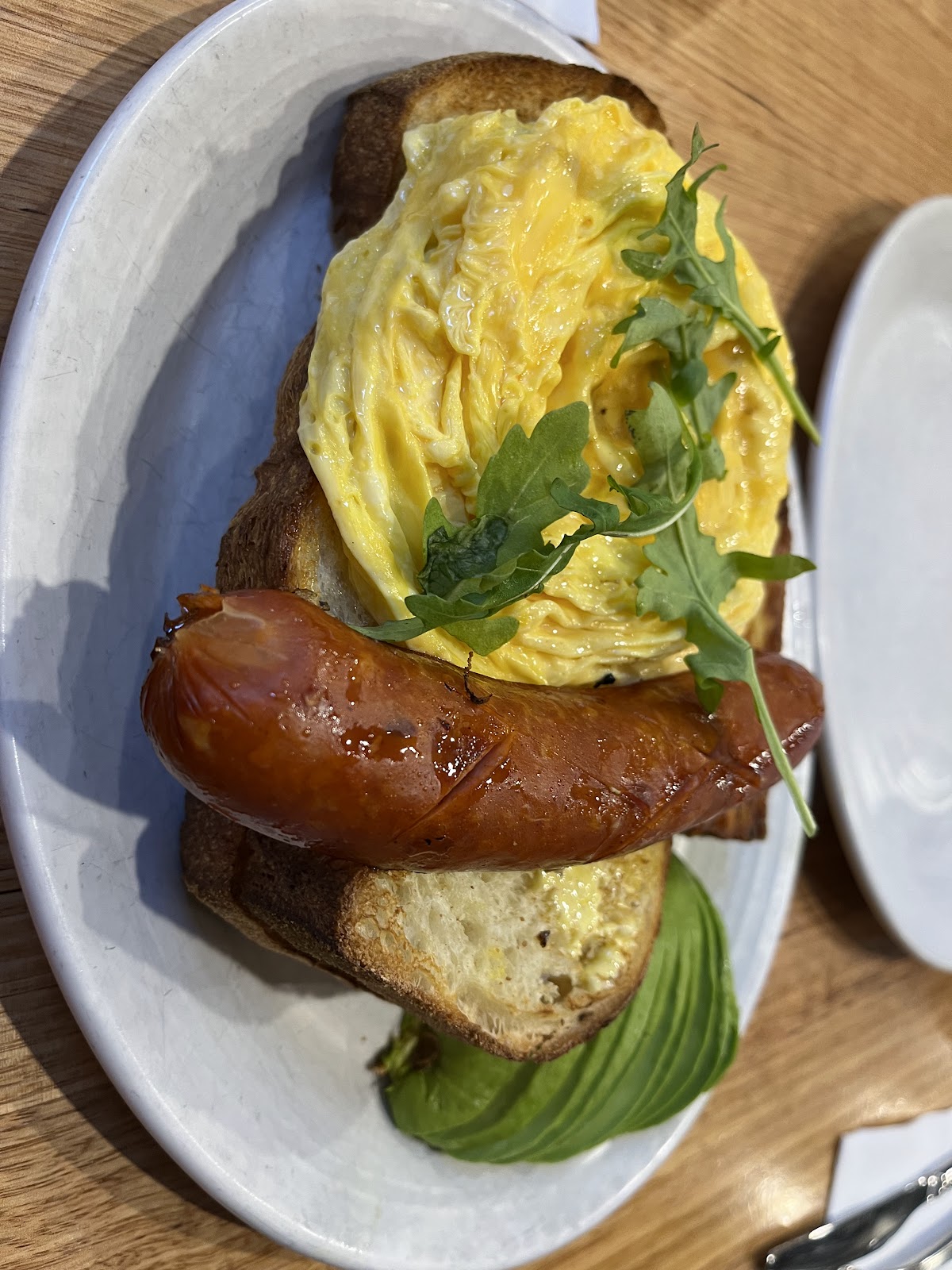 A toasted sourdough sandwich with scrambled egg, sausage, avocado and rocket, on a white plate at Salt n Pepa Cafe.